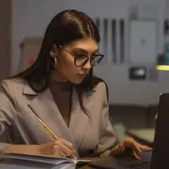 Kobieta w okularach pracuje przy laptopie i notuje. Zastanawia się, ile nadgodzin w roku jest w stanie przepracować.