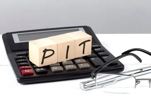 Drewniane klocki z napisem PIT leżą na kalkulatorze. Obok okulary i długopis. Symbol niepełnosprawności 12-c ulgi może być związany z rozliczeniem podatkowym.