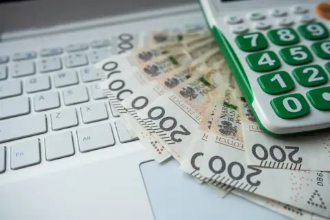 Kalkulator i banknoty 200 zł na klawiaturze laptopa. Obrazek sugeruje analizę finansową, być może związaną z grupami podatkowymi.