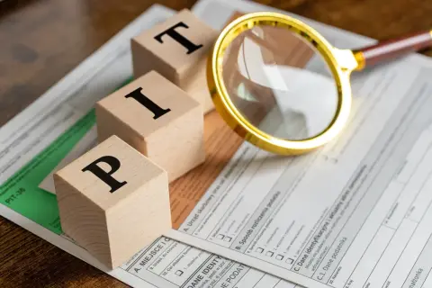 Drewniane klocki z literami P, I, T leżą na formularzu PIT. Obok znajduje się złota lupa, sugerująca analizę podatku dochodowego.