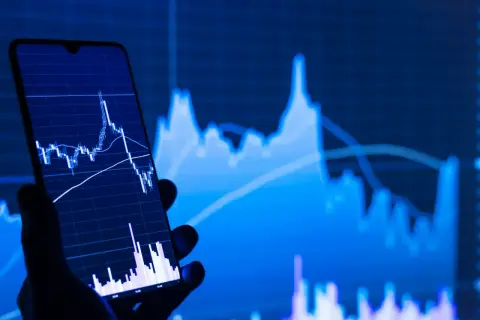 Dłoń trzyma smartfon z wykresem świecowym, na tle większego wykresu. Analiza wskaźnikowa w akcji.