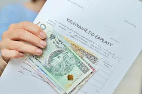 Dłoń trzyma wezwanie do zapłaty i polskie banknoty. Czy to odpowiedź na pytanie, ile zarabia komornik?
