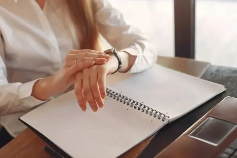 Kobieta w białej koszuli sprawdza zegarek, pracując po godzinach. Na biurku leży otwarty notatnik.
