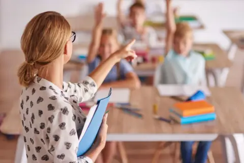 Nauczycielka w klasie z uczniami podnoszącymi ręce. Pokazuje, jak zostać nauczycielem przedmiotów zawodowych.