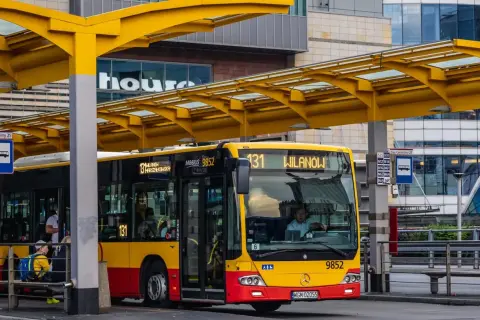 Kierowca autobusu linii 131 dojeżdża do przystanku Wilanów. Żółto-czerwony pojazd czeka na pasażerów pod żółtą wiatą.