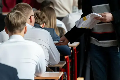 Nauczyciel rozdaje arkusze egzaminacyjne uczniom siedzącym w ławkach.