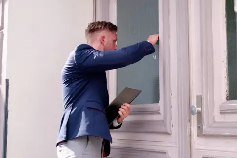 Mężczyzna w garniturze puka do drzwi, trzymając teczkę. Czy to wizyta komornika? Zastanawiasz się, ile może zabrać komornik?