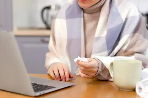 Osoba owinięta kocem, z chusteczką w dłoni, pracuje przy laptopie. Obok kubek i zużyte chusteczki. Może to być sytuacja związana z art. 148 kp.