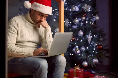 Mężczyzna w czapce Mikołaja siedzi przy choince, pracując na laptopie. Widać prezenty pod choinką. To smutne **święta**.