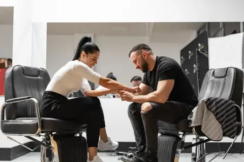 Barber w czarnym t-shircie i podartych jeansach siedzi na fotelu, trzymając dłoń klientki. Zastanawia się, ile zarabia barber.