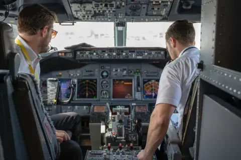Dwóch pilotów samolotu w kokpicie. Jeden z nich, pilot, patrzy na instrumenty, drugi obsługuje sterowanie.
