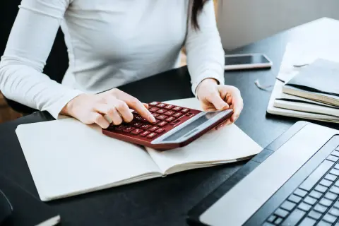 Kobieta liczy wydatki na własne cele mieszkaniowe, przygotowując PIT-39. Na biurku kalkulator, notatnik i laptop.