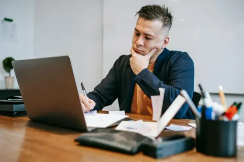 Zamyślony audytor siedzi przy biurku, pisząc coś na papierze i patrząc na ekran laptopa.