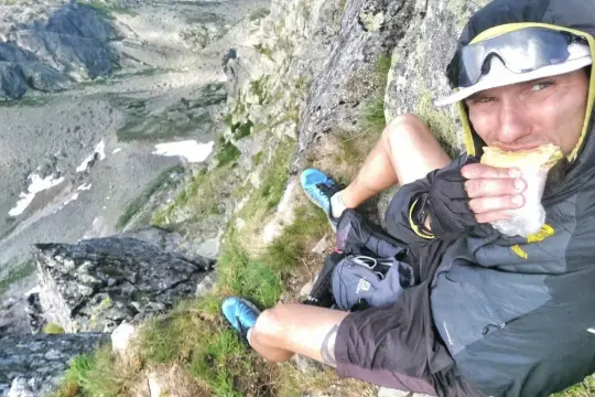 Mężczyzna w okularach przeciwsłonecznych i kapturze siedzi na skale, jedząc kanapkę. Widok na góry. To jego pierwsza górska wyprawa, praca bez doświadczenia, ale daje radę!