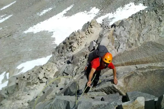 Alpinista w pomarańczowym kasku wspina się po skalistym zboczu z plecakiem.