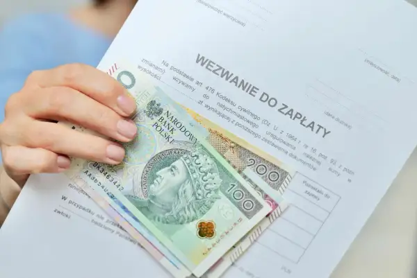 Dłoń trzyma wezwanie do zapłaty i polskie banknoty. Czy to odpowiedź na pytanie, ile zarabia komornik?