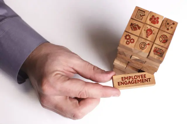Dłoń wyciąga klocek z napisem "EMPLOYEE ENGAGEMENT", budując wieżę symbolizującą zaangażowanie pracowników i sukces firmy.